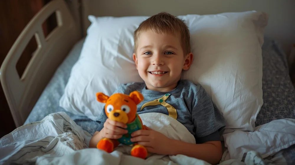 A young patient smiling in his hospital bed with a stuffed animal